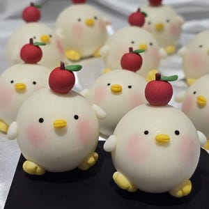 May include: A collection of round, white chick-shaped pastries with rosy cheeks, yellow beaks and feet, and a red apple on top. The pastries are arranged on a black surface, with a white, textured background.