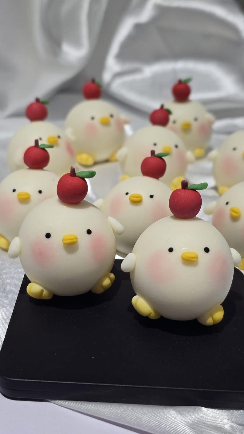 May include: A collection of round, white chick-shaped pastries with rosy cheeks, yellow beaks and feet, and a red apple on top. The pastries are arranged on a black surface, with a white, textured background.