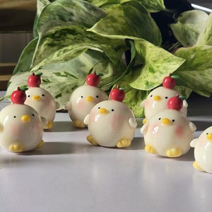 May include: A collection of ceramic chick figurines, each with a red apple on its head. The chicks have a cream-colored body, yellow feet, and a pink blush on their cheeks. The figurines are arranged on a white surface.
