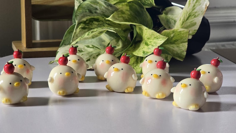 May include: A collection of ceramic chick figurines, each with a red apple on its head. The chicks have a cream-colored body, yellow feet, and a pink blush on their cheeks. The figurines are arranged on a white surface.