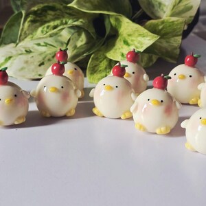 May include: A collection of ceramic chick figurines, each with a red apple on its head. The chicks are white with yellow feet and beaks, and pink cheeks. The figurines are arranged on a white surface.