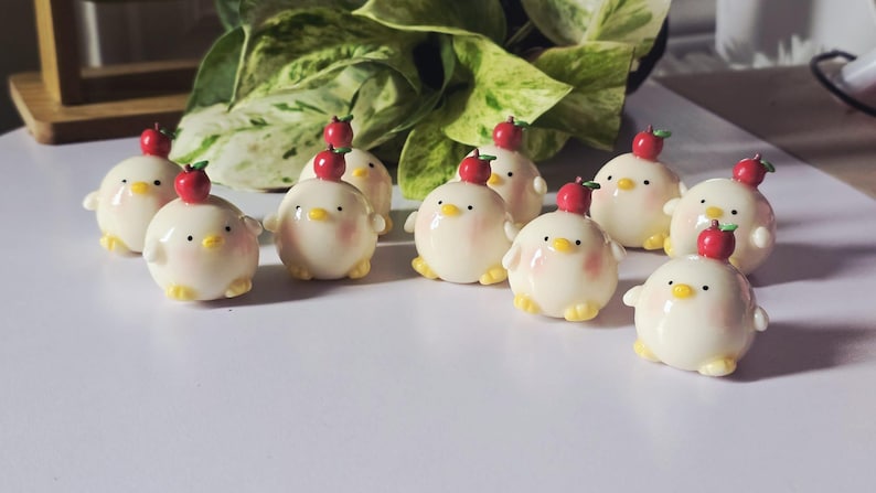 May include: A collection of ceramic chick figurines, each with a red apple on its head. The chicks are white with yellow feet and beaks, and pink cheeks. The figurines are arranged on a white surface.