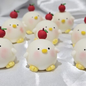 May include: A collection of adorable, round, white chick-shaped treats with rosy cheeks, yellow feet, and small black eyes. Each chick has a red apple with a green stem on its head. The treats are arranged on a white surface.