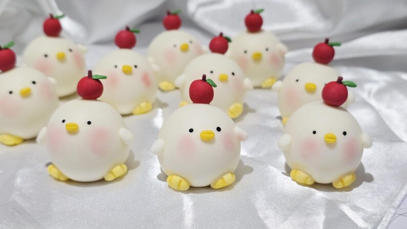 May include: A collection of adorable, round, white chick-shaped treats with rosy cheeks, yellow feet, and small black eyes. Each chick has a red apple with a green stem on its head. The treats are arranged on a white surface.