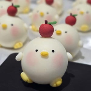 May include: A collection of adorable, round, white chick-shaped pastries, each with a red apple on top. The chicks have rosy cheeks, yellow beaks and feet, and small black eyes. They are arranged on a black surface.