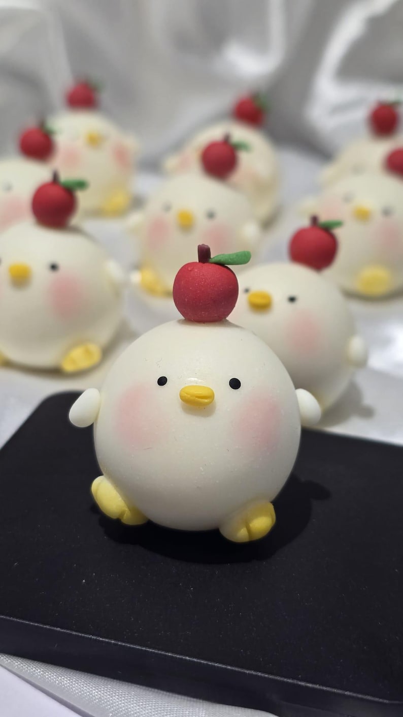 May include: A collection of adorable, round, white chick-shaped pastries, each with a red apple on top. The chicks have rosy cheeks, yellow beaks and feet, and small black eyes. They are arranged on a black surface.