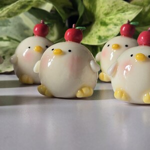 May include: A collection of adorable, round, cream-colored chick figurines with yellow feet and beaks, and rosy cheeks. Each chick has a red apple on its head. The figurines are arranged on a white surface.
