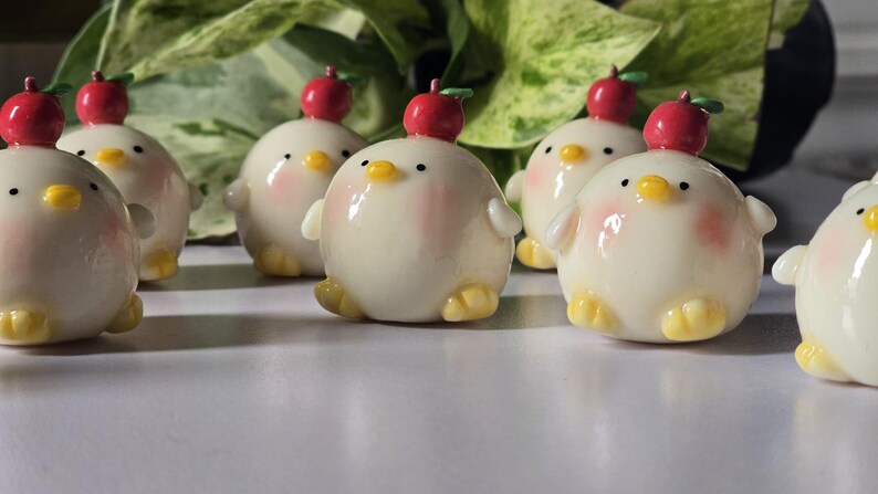 May include: A collection of adorable, round, cream-colored chick figurines with yellow feet and beaks, and rosy cheeks. Each chick has a red apple on its head. The figurines are arranged on a white surface.