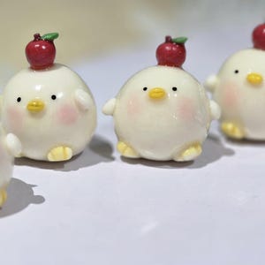 May include: Four adorable, round, white chick figurines with rosy cheeks, yellow feet, and small wings. Each chick has a red apple with a green leaf on its head. The figurines are arranged on a white surface.