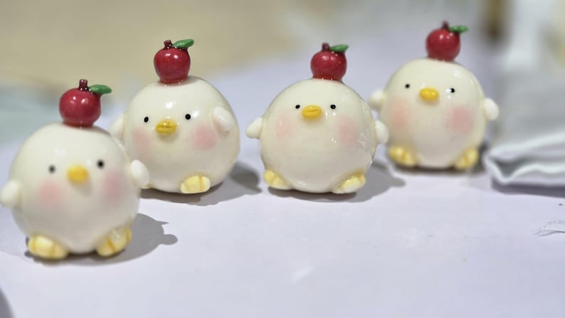 May include: Four adorable, round, white chick figurines with rosy cheeks, yellow feet, and small wings. Each chick has a red apple with a green leaf on its head. The figurines are arranged on a white surface.