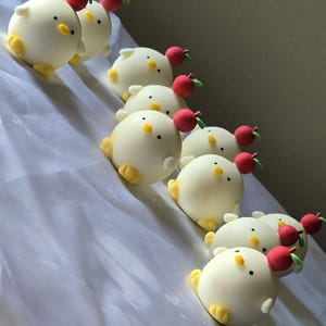 May include: A collection of white, round chick-shaped treats with yellow feet and beaks, and small red apple decorations on top. The treats are arranged in a diagonal line on a white surface.