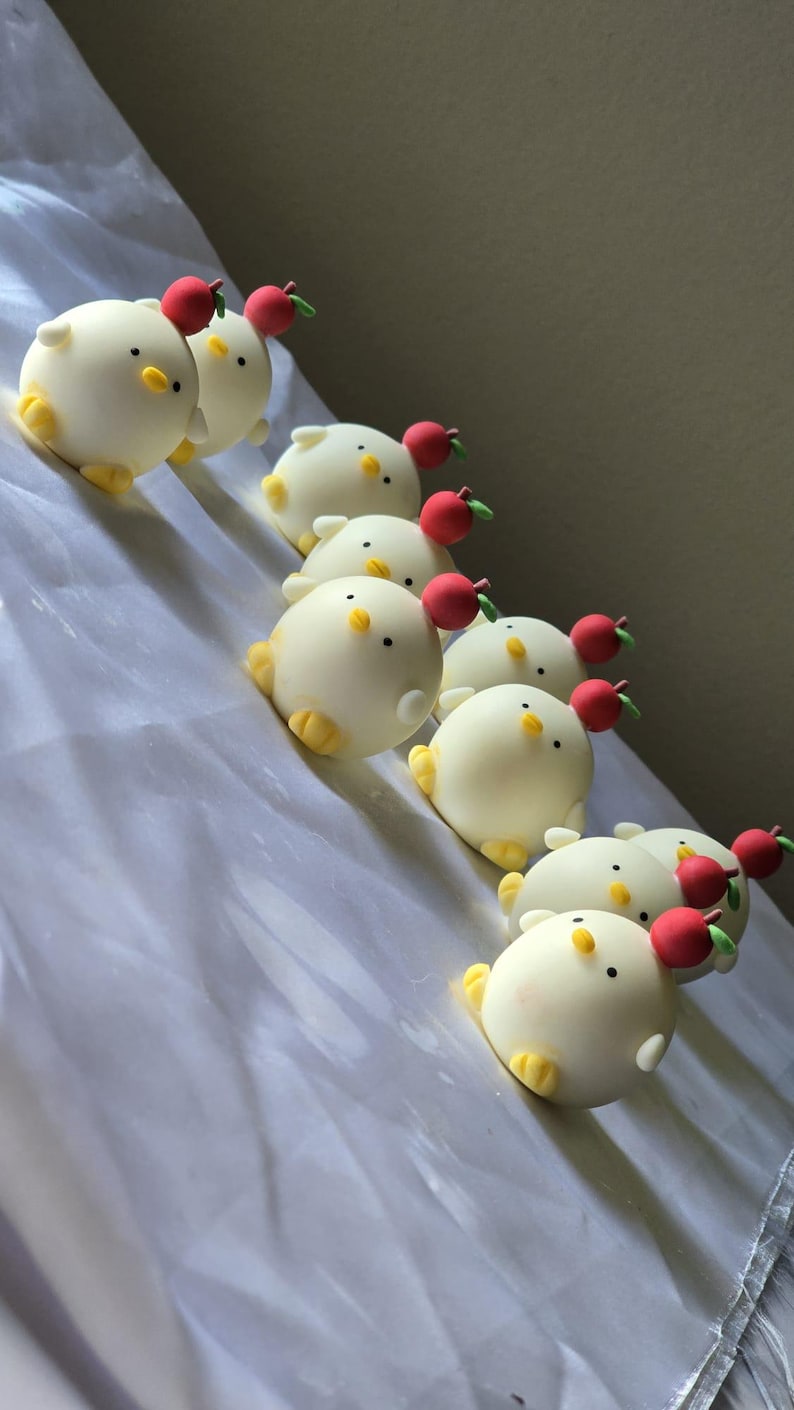 May include: A collection of white, round chick-shaped treats with yellow feet and beaks, and small red apple decorations on top. The treats are arranged in a diagonal line on a white surface.