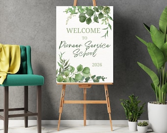May include: A rectangular white sign on a wooden easel featuring green watercolor eucalyptus leaves at the top and bottom. The text reads WELCOME TO Pioneer Service School 2026 in a mix of dark green serif and script fonts on a textured surface.