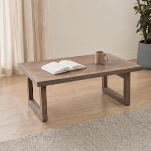 May include: A low rectangular coffee table made of brown wood with a visible grain texture. The table features four sturdy legs connected by horizontal base supports. An open white book and a solid brown ceramic mug sit on the tabletop surface.
