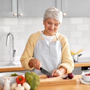 K&ouml;nnte beinhalten: Eine Frau in einem gelben Pullover und einer gestreiften Sch&uuml;rze schneidet rote Zwiebeln auf einem Holzschneidebrett in einer K&uuml;che. Verschiedenes Gem&uuml;se und eine Salatsch&uuml;ssel stehen auf der Theke. Eine &Ouml;lflasche und eine Sch&uuml;ssel mit Tomaten sind ebenfalls zu sehen.