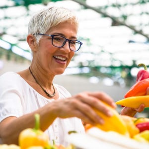 K&ouml;nnte beinhalten: Eine Frau l&auml;chelt, w&auml;hrend sie auf einem Bauernmarkt bunte Paprika ausw&auml;hlt. Die Paprika sind rot, orange und gelb. Sie tr&auml;gt eine Brille und ein wei&szlig;es Oberteil mit einer Halskette.