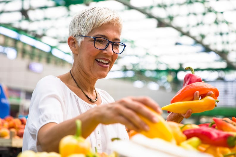 K&ouml;nnte beinhalten: Eine Frau l&auml;chelt, w&auml;hrend sie auf einem Bauernmarkt bunte Paprika ausw&auml;hlt. Die Paprika sind rot, orange und gelb. Sie tr&auml;gt eine Brille und ein wei&szlig;es Oberteil mit einer Halskette.