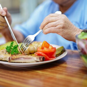 K&ouml;nnte beinhalten: Ein Teller mit Essen mit einem Schweinekotelett, Tomate und Essiggurke wird mit Messer und Gabel geschnitten. Eine Sch&uuml;ssel Salat ist im Hintergrund. Das Essen steht auf einem Holztisch.