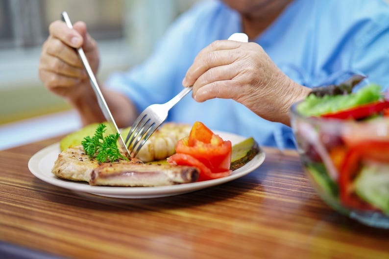 K&ouml;nnte beinhalten: Ein Teller mit Essen mit einem Schweinekotelett, Tomate und Essiggurke wird mit Messer und Gabel geschnitten. Eine Sch&uuml;ssel Salat ist im Hintergrund. Das Essen steht auf einem Holztisch.