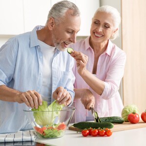 K&ouml;nnte beinhalten: Ein Paar bereitet in einer hellen K&uuml;che einen Salat zu. Eine Person gibt Salat in eine klare Glassch&uuml;ssel mit Tomaten und anderem Gem&uuml;se. Die andere Person schneidet eine Gurke. Frische Tomaten, Gurken und Salat sind sichtbar.