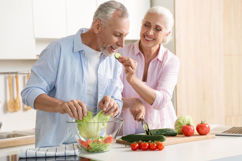 K&ouml;nnte beinhalten: Ein Paar bereitet in einer hellen K&uuml;che einen Salat zu. Eine Person gibt Salat in eine klare Glassch&uuml;ssel mit Tomaten und anderem Gem&uuml;se. Die andere Person schneidet eine Gurke. Frische Tomaten, Gurken und Salat sind sichtbar.