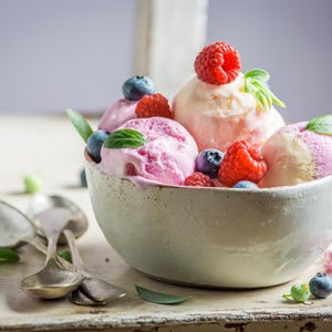 K&ouml;nnte beinhalten: Eine Keramikschale gef&uuml;llt mit Kugeln aus rosa und pfirsichfarbenem Eis, garniert mit frischen Himbeeren, Blaubeeren und Minzbl&auml;ttern. Silberl&ouml;ffel liegen neben der Schale auf einer rustikalen Holzoberfl&auml;che.