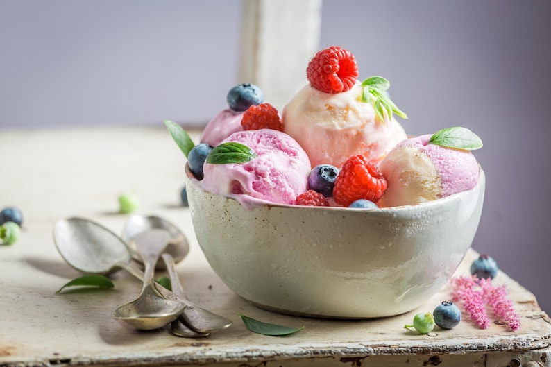 K&ouml;nnte beinhalten: Eine Keramikschale gef&uuml;llt mit Kugeln aus rosa und pfirsichfarbenem Eis, garniert mit frischen Himbeeren, Blaubeeren und Minzbl&auml;ttern. Silberl&ouml;ffel liegen neben der Schale auf einer rustikalen Holzoberfl&auml;che.