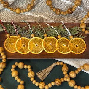 May include: Six dried orange slice ornaments with twine hangers are arranged on a wooden tray with greenery. Wooden bead garlands and a jute tassel are also in the image, suggesting a rustic, natural theme.