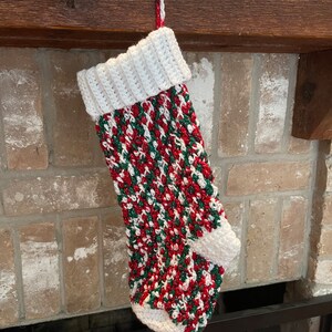 May include: A white, red, and green crocheted Christmas stocking with a white cuff hanging from a fireplace mantel.