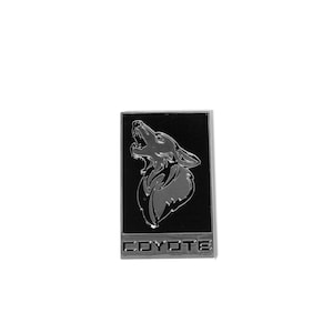 May include: A silver metal emblem with a black background featuring a howling coyote silhouette and the word "COYOTE" in silver lettering.