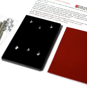 May include: Close-up of two black metal plates with four holes each, and a red metal plate. The plates are used to install a car badge. Instructions for installation are visible in the background. The instructions mention using 10mm nuts, a deep socket, and a 7mm deep socket.