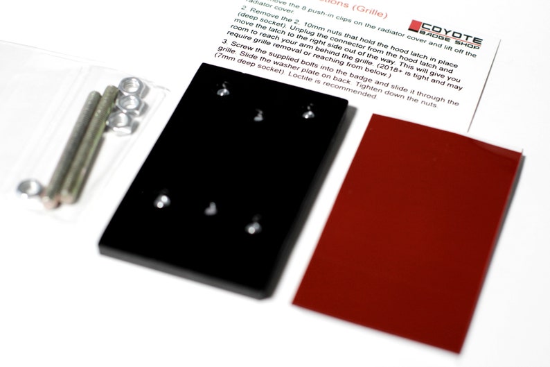May include: Close-up of two black metal plates with four holes each, and a red metal plate. The plates are used to install a car badge. Instructions for installation are visible in the background. The instructions mention using 10mm nuts, a deep socket, and a 7mm deep socket.