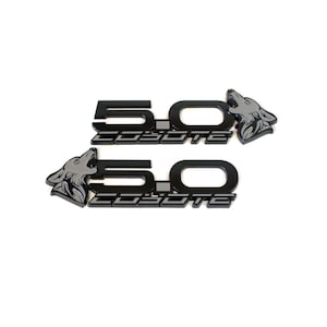 May include: Two black 5.0 Coyote emblems with a silver coyote head graphic. The emblems are for a car or truck.