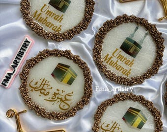 May include: Four round white coasters with gold glitter edges. The coasters feature a gold image of the Kaaba, a cube-shaped building in Mecca, Saudi Arabia. The text "Umrah Mubarak" is written in gold on the coasters. The coasters are on a white surface with gold metal stands.