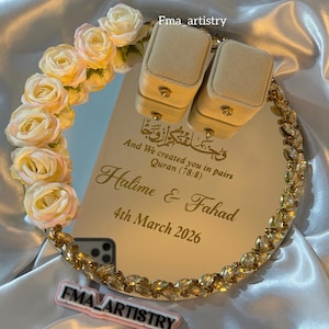 Puede incluir: Una bandeja de boda con espejo, borde dorado con joyas y adornos florales. La bandeja muestra dos cajas para anillos y los nombres "Halime & Fahad" con la fecha "4 de marzo de 2026". También se ve el texto "And We created you in pairs".
