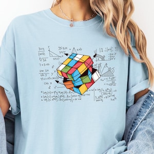 May include: A light blue t-shirt with a colorful Rubik's Cube graphic and mathematical equations printed on the front. The shirt features a casual, relaxed fit.