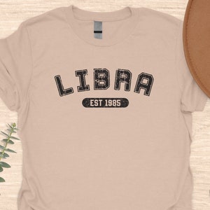 May include: A beige t-shirt with a black graphic that says "LIBRA EST 1985" in a vintage collegiate style font.
