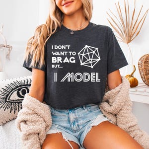May include: A dark gray t-shirt with white text that reads "I don't want to brag but... I model" and a geometric shape graphic.