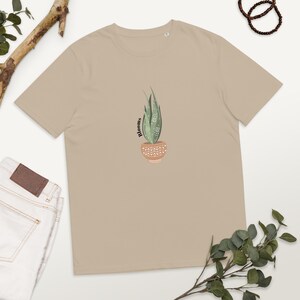 Puede incluir: Una camiseta beige con una planta verde en una maceta marrón y la palabra "blooms" escrita verticalmente en el lado izquierdo de la maceta.