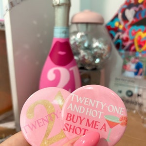 May include: Two pink button badges with gold glitter accents. One badge says "Twenty-one and hot buy me a shot" with a pink cocktail glass and orange slice. The other badge says "Twenty-one" with a large gold glitter "21".