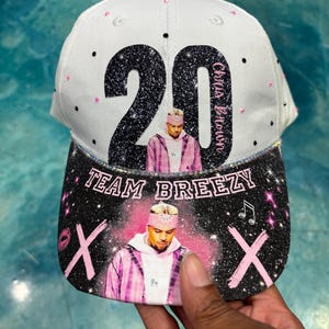 Könnte beinhalten: Weiß-schwarze Baseballkappe mit der Zahl 20 in Glitzer und dem Namen "Chris Brown". Der Schirm zeigt den Text "TEAM BREEZY" und ein Bild von Chris Brown.