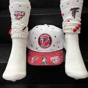 Op de afbeelding: Witte Atlanta Falcons baseballpet en bijpassende witte sokken. De pet heeft het teamlogo en de woorden "Atlanta Falcons". De sokken hebben de woorden "Rise Up" en het teamlogo. Rode en zwarte accenten.