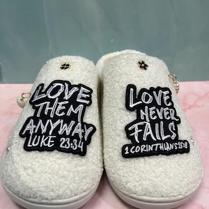 Puede incluir: Un par de zapatillas blancas y esponjosas con parches negros. Uno dice "LOVE THEM ANYWAY LUKE 23:34", el otro "LOVE NEVER FAILS 1 CORINTHIANS 13:8". Las zapatillas tienen detalles dorados.