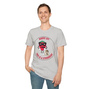 May include: A light grey t-shirt with a cartoon strawberry wearing a mask and holding a money bag. The text "HANDS UP! THIS IS A STROBBERY" is printed around the strawberry in a circular design.