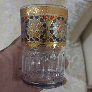 May include: A clear glass drinking glass with a gold and blue geometric pattern around the top. The glass has a textured surface.