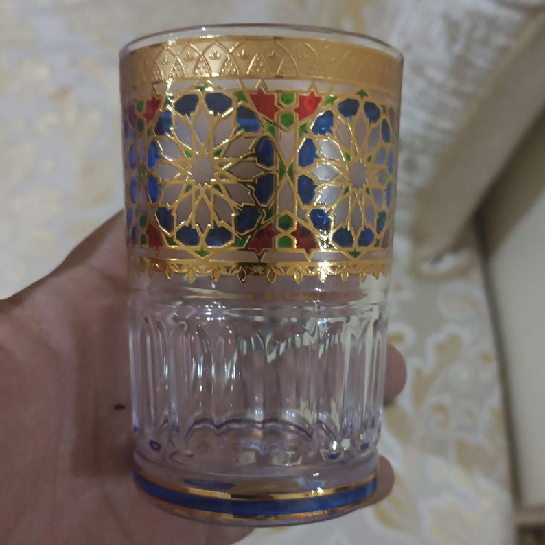 May include: A clear glass drinking glass with a gold and blue geometric pattern around the top. The glass has a textured surface.