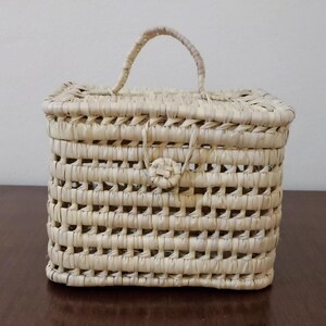 May include: A small, rectangular woven basket with a handle. The basket is made of natural straw and has a light beige color.