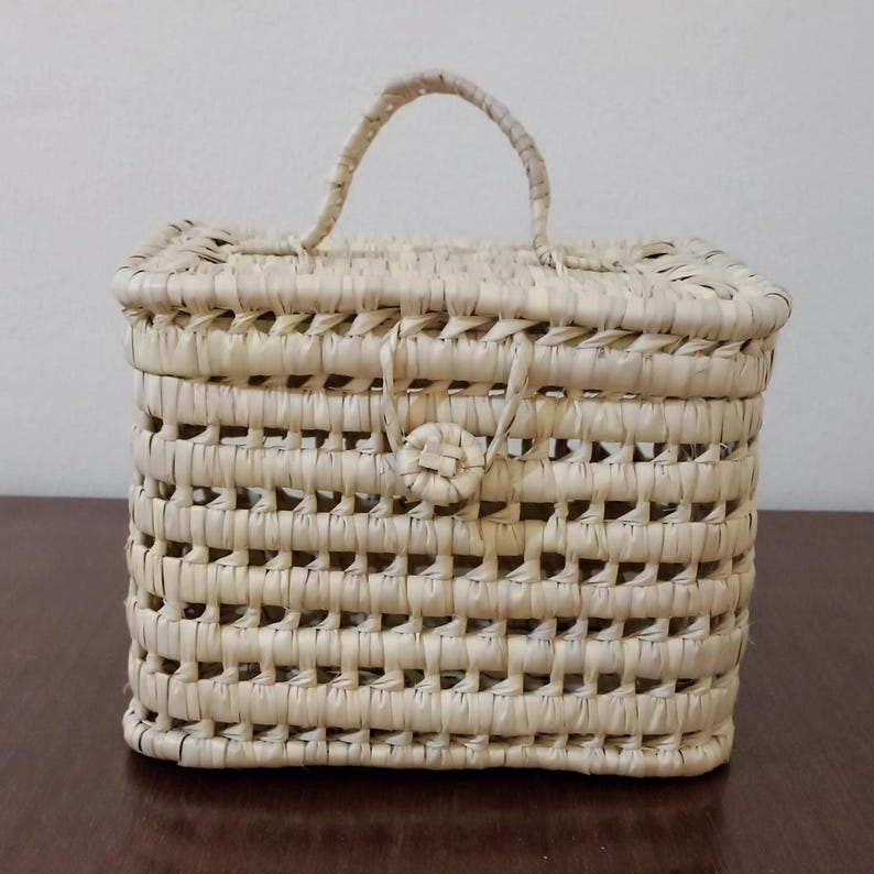 May include: A small, rectangular woven basket with a handle. The basket is made of natural straw and has a light beige color.