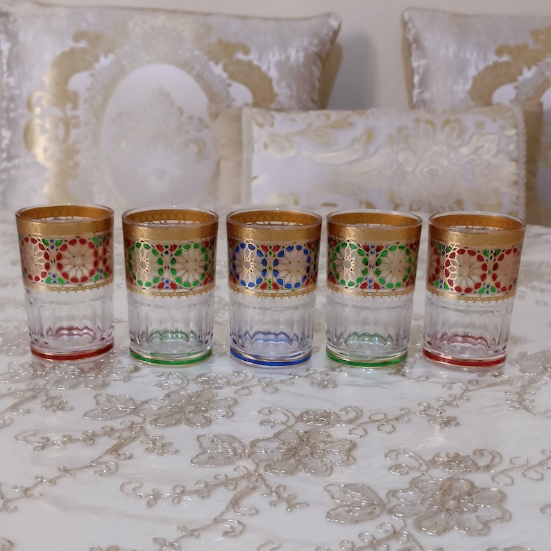 May include: Five clear glass tumblers with gold, red, green, and blue geometric patterns around the top rim. The glasses are lined up in a row on a white tablecloth.