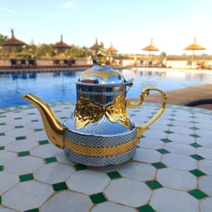 Könnte beinhalten: Eine silberne und goldene Teekanne mit einem dekorativen Design steht auf einer weißen und grünen gefliesten Oberfläche. Die Teekanne hat einen runden Körper und einen Ausguss an der Seite. Der Griff ist gebogen und an der Oberseite der Teekanne befestigt.